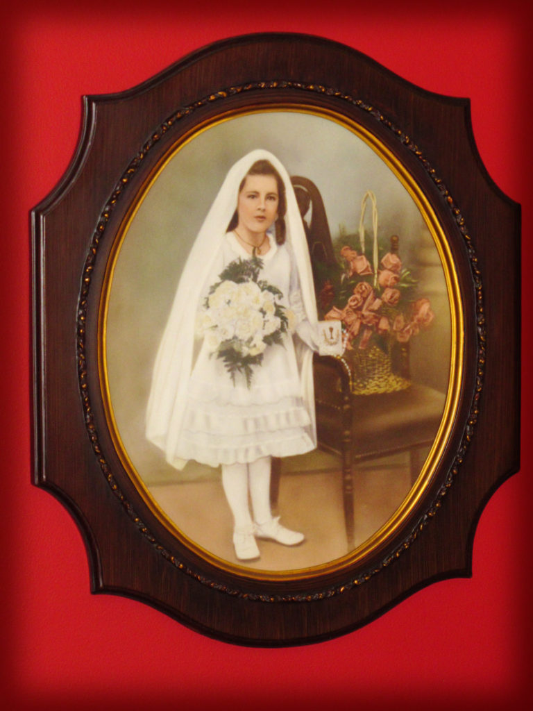 Photo of a antique 100 year old photo of a young girl in her Communion Dress standing next to a chair with pink flowers on it. Girl holds flowers and a prayer book.